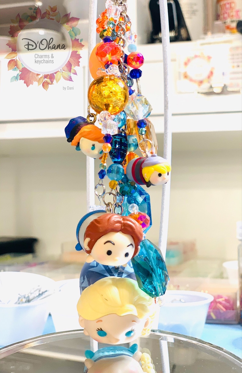 May include: A colorful charm keychain featuring characters from the animated film Frozen. The keychain includes a blue and white charm with a character from Frozen, a blue and white charm with a character from Frozen, a blue and white charm with a character from Frozen, and a blue and white charm with a character from Frozen. The keychain is adorned with blue, white, and gold beads.
