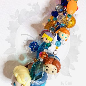 May include: A colorful charm bracelet featuring a variety of beads, including clear, blue, orange, and green. The bracelet also includes small figurines of characters from the movie Frozen, including Elsa, Anna, Kristoff, and Olaf. The bracelet is finished with a silver charm that reads "by Dani".