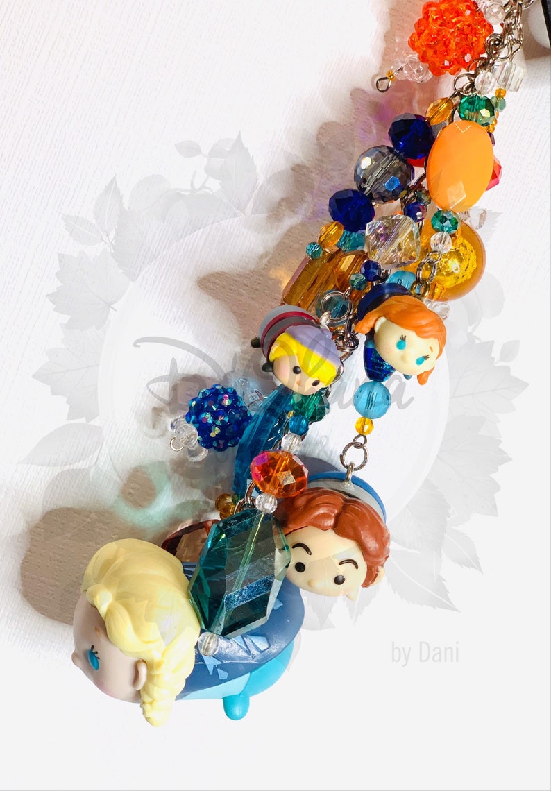 May include: A colorful charm bracelet featuring a variety of beads, including clear, blue, orange, and green. The bracelet also includes small figurines of characters from the movie Frozen, including Elsa, Anna, Kristoff, and Olaf. The bracelet is finished with a silver charm that reads "by Dani".
