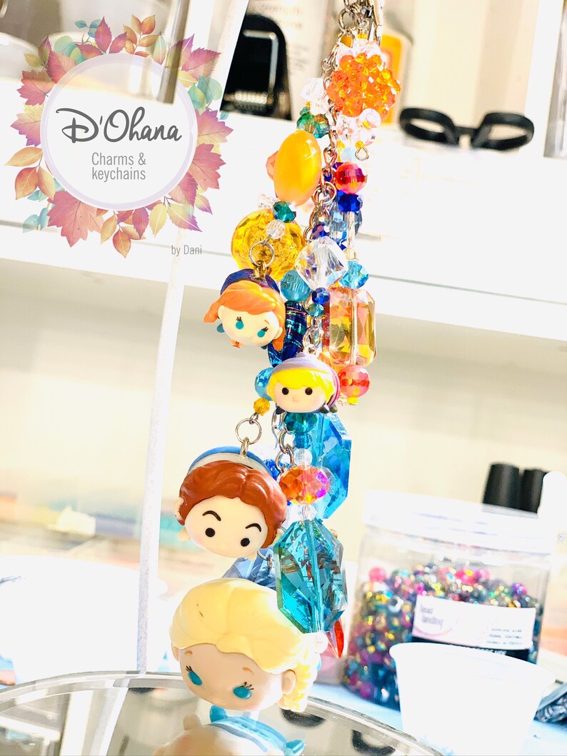 May include: A colorful charm bracelet featuring a variety of beads, including clear crystals, orange, yellow, and blue beads. The bracelet also features four small, cartoon-style character charms. The bracelet is labeled "D'Ohana Charms & Keychains by Dani."