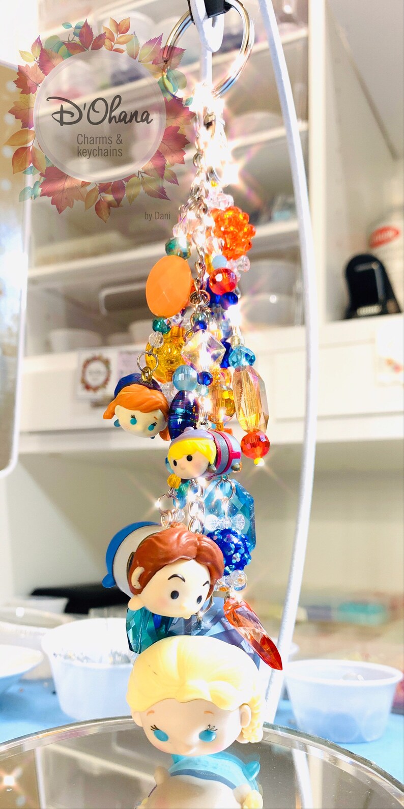 May include: A colorful keychain featuring a variety of charms, including small figurines of characters from the Disney movie Frozen. The keychain is made with beads, crystals, and metal accents. The keychain is labeled "D'Ohana Charms & Keychains by Dani."