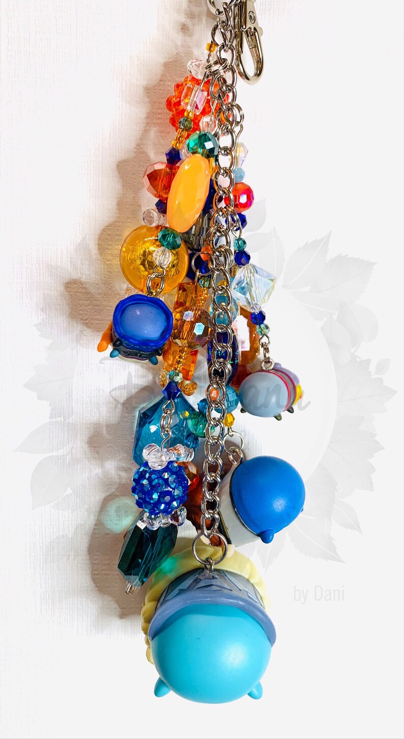 May include: A colorful charm keychain with a blue, yellow, and white character charm. The keychain features a variety of beads, including clear crystals, orange, blue, and green beads, and a silver chain.