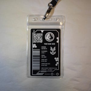 Könnte beinhalten: Ein schwarz-weißes ID-Abzeichen mit dem Text "YOUR NAME HERE" und anderen Details. Das Abzeichen befindet sich in einer transparenten Kunststoffhalterung mit einem angebrachten Schlüsselband. Das Abzeichen enthält einen QR-Code und das UNSC-Logo.