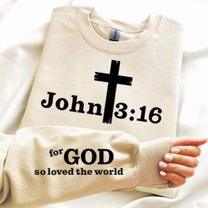 May include: A beige sweatshirt with the text "John 3:16" and a black cross graphic. The sleeve displays the text "for GOD so loved the world" in black. The sweatshirt is made of a soft material.