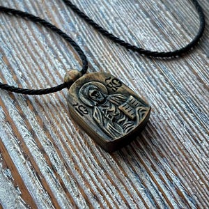 May include: A wooden pendant necklace featuring a carved religious icon. The pendant is rectangular with rounded edges and is suspended from a black cord. The icon depicts a figure with a halo and cross. The background is a wooden surface.