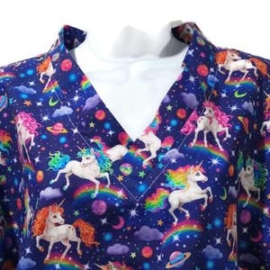 Peut inclure: Un haut de blouse bleu foncé avec un col en V, orné d'un motif d'unicorns blancs avec des crinières et des queues arc-en-ciel, des arcs-en-ciel, des planètes, des étoiles et des nuages. Le haut de blouse a deux poches.