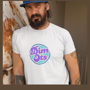 Peut inclure: Un t-shirt blanc avec un motif circulaire portant l'inscription "Dim Ots" en violet et turquoise. Le t-shirt est porté par une personne portant une casquette de baseball bleue. La personne a une barbe et des tatouages sur le bras.