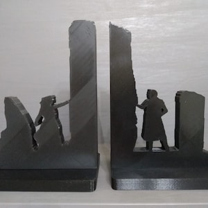 Könnte beinhalten: Ein Paar schwarze Buchstützen mit einem Silhouettendesign von Figuren, die mit einer bröckelnden Wand interagieren. Die Buchstützen bestehen aus einem soliden Material und haben eine rechteckige Basis für Stabilität. Ein einzigartiges dekoratives Element für jedes Bücherregal.