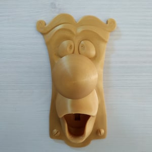 Op de afbeelding: Een 3D-geprinte, wandgemonteerde flessenopener in de vorm van een cartoon gezicht met een grote neus en een open mond. De flessenopener is in een lichtbruine kleur geverfd.