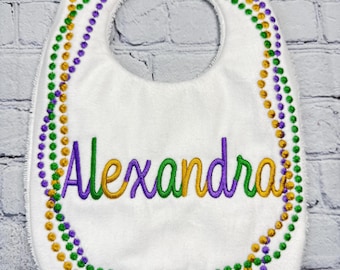 May include: White baby bib with a green, purple, and gold beaded border. The name "Alexandra" is embroidered in the center in green, purple, and gold thread.