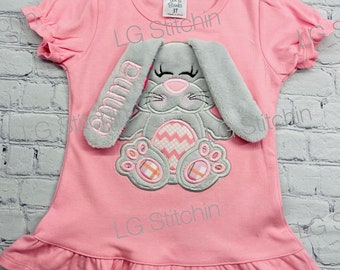 May include: Pink short-sleeved shirt with a gray bunny applique and the name "Emma" embroidered on the bunny's ear. The bunny is holding a pink and white Easter egg.