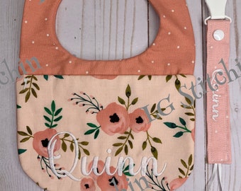May include: A pink and white floral baby bib with the name "Quinn" embroidered on it. The bib has a snap closure and comes with a matching pink and white floral strap.