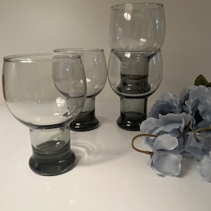 May include: Four gray-tinted glass goblets with rounded bowls and short, dark gray stems. One goblet is stacked on top of another. A cluster of blue artificial flowers is in the background. The glasses are on a white surface.