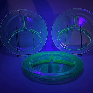 May include: Three clear glass divided plates with a neon green glow under a black light. The plates are round with three sections. One stack of plates is on the bottom.