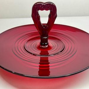 May include: A red glass serving tray with a heart-shaped handle. The tray has a concentric circle design.