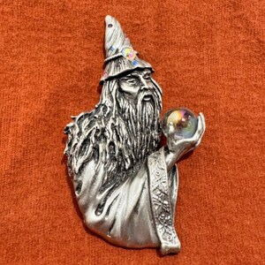 May include: A silver-toned metal brooch depicting a bearded wizard wearing a pointed hat adorned with small, colorful iridescent stones. The wizard holds a clear, spherical orb in one hand, and the robe features intricate floral detailing.