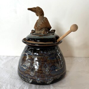 May include: A ceramic honey pot with a bird-shaped lid and a wooden dipper. The pot has a dark brown glaze with blue and white accents.