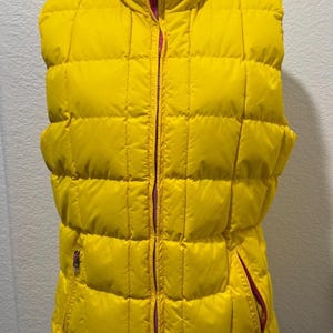 May include: A bright yellow puffer vest with a high collar and a full-length zipper. The vest has a quilted design and two side pockets with zippers. The interior lining is a contrasting pink color.