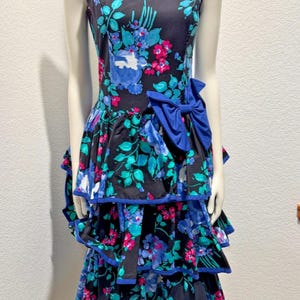 May include: A sleeveless floral dress with a tiered skirt and a large blue bow. The dress features a black background with blue, teal, and pink floral patterns. The edges are trimmed with blue piping.