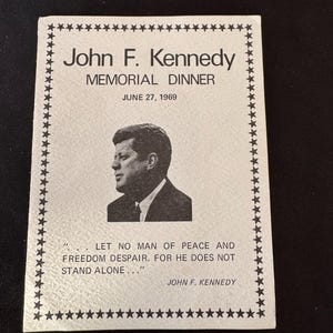 May include: A black and white photo of John F. Kennedy on a white background with a black border. The text reads "John F. Kennedy Memorial Dinner June 27, 1969" and "LET NO MAN OF PEACE AND FREEDOM DESPAIR. FOR HE DOES NOT STAND ALONE..."  "JOHN F. KENNEDY"