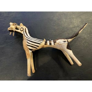 May include: A handcrafted, stylized animal figure with a zebra-striped body and a long tail. The figure has a light-colored body with black stripes and accents, and a fur-like mane. The legs are thin and stick-like, and the figure is posed in a standing position.
