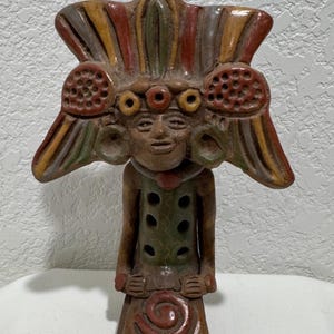 May include: A hand-painted ceramic figurine depicting a stylized figure with a large headdress. The figure is adorned with multiple colors, including red, yellow, green, and brown. The figurine has a detailed face and is standing upright.