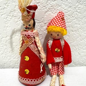 May include: Two vintage Christmas ornaments. One is a red and cream-colored figure with a tall hat and a decorative collar. The other is a red and white polka-dotted figure with a matching hat and shorts. Both have yellow hair.