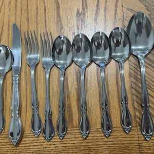 May include: A collection of stainless steel silverware, including spoons, forks, and knives, arranged on a wooden surface. The silverware has a decorative handle design. The spoons vary in size, and the forks have four tines.