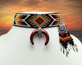 May include: A beaded choker necklace with a geometric pattern in black, white, yellow, and red. It features a crescent-shaped pendant and a dangling earring with beads and silver-colored charms. The jewelry is displayed on a white mannequin head.