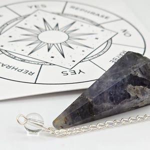 Peut inclure: Un pendule en cristal violet foncé à facettes attaché à une chaîne en métal argenté avec une petite perle transparente à l'extrémité. Derrière le pendule se trouve un tableau de divination circulaire blanc avec un soleil central et les mots YES, NO, REPHRASE.