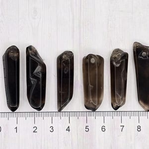 May include: Six dark brown crystal pendants of varying shapes and sizes are arranged on a white surface next to a ruler. Each pendant has a hole drilled at the top. The ruler shows measurements in inches.
