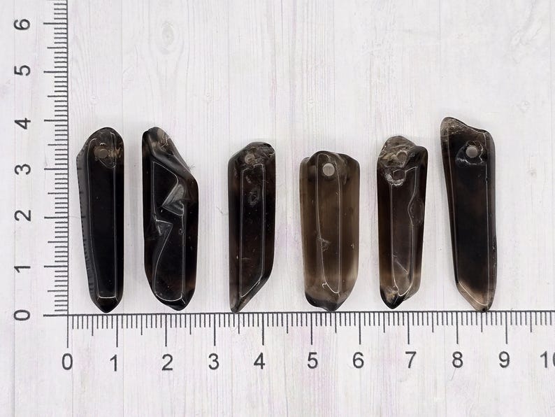 May include: Six dark brown crystal pendants of varying shapes and sizes are arranged on a white surface next to a ruler. Each pendant has a hole drilled at the top. The ruler shows measurements in inches.