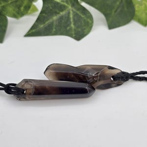 May include: Two smoky quartz crystal pendants with black cords. The crystals are a translucent brown color and are cut into a hexagonal shape. The pendants are displayed on a white surface with green leaves in the background.