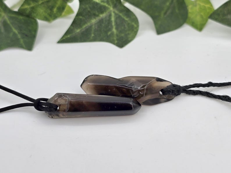 May include: Two smoky quartz crystal pendants with black cords. The crystals are a translucent brown color and are cut into a hexagonal shape. The pendants are displayed on a white surface with green leaves in the background.