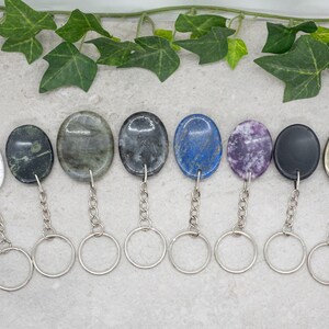 Op de afbeelding: Acht verschillende ovale edelsteen sleutelhangers met zilveren sleutelringen. De stenen zijn wit, zwart, groen, grijs, blauw, paars, zwart en goud.