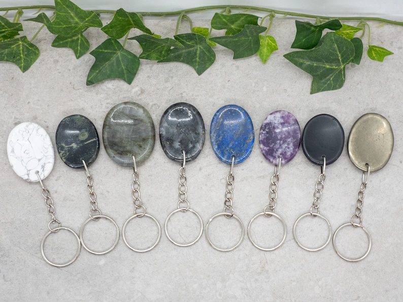 Op de afbeelding: Acht verschillende ovale edelsteen sleutelhangers met zilveren sleutelringen. De stenen zijn wit, zwart, groen, grijs, blauw, paars, zwart en goud.