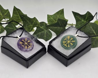 May include: Two round stone pendants on black cords, one purple amethyst and one green aventurine, each featuring a gold Vegvisir symbol. The pendants are displayed in individual black gift boxes with white padding.