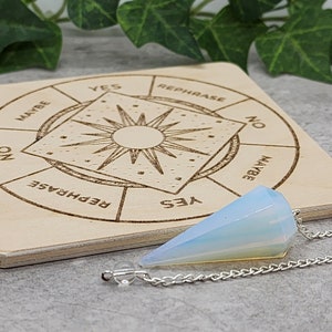 Puede incluir: Un tablero de péndulo de madera con las palabras "Sí", "No", "Tal vez" y "Reformular" impresas alrededor del borde. Un péndulo de ópalo blanco cuelga de una cadena plateada.