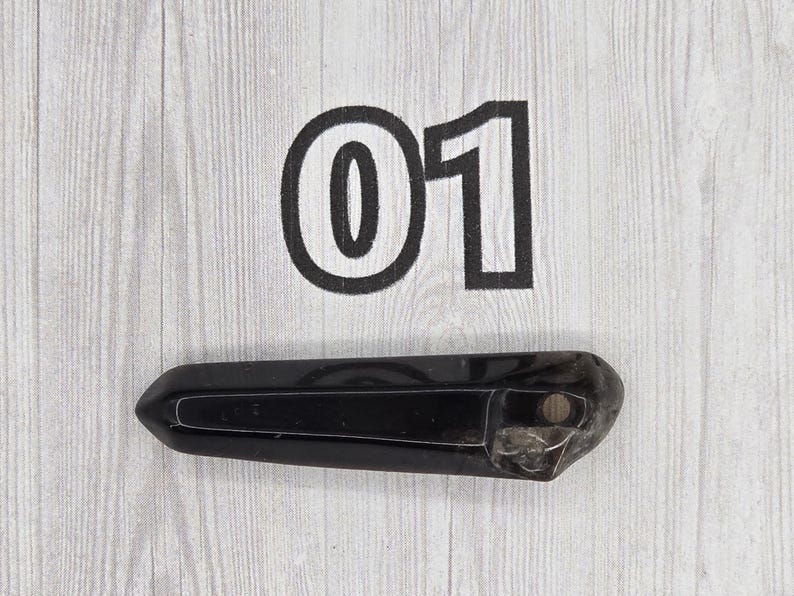 May include: A black, elongated crystal with a polished surface. The number "01" is printed in black above the crystal on a light wood-grain background. The crystal is approximately 5 inches long.