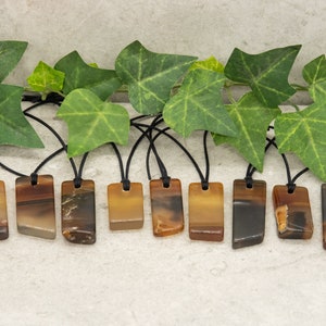 May include: Ten brown and black agate stone pendants on black cords. The pendants are rectangular and have a natural, polished finish.