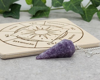 Pode incluir: Um pêndulo de ametista roxo com uma corrente de prata pendurada sobre uma tábua de madeira com um design circular e as palavras "SIM", "NÃO", "TALVEZ" e "REFRASEAR" escritas ao redor do círculo.