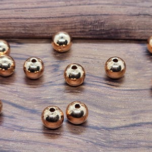 Peut inclure: Une collection de onze perles sphériques avec une finition métallique en or rose poli. Chaque perle présente une surface lisse et réfléchissante ainsi qu'un trou central unique pour l'enfilage, disposées de manière éparse.