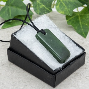 Könnte beinhalten: Eine dunkelgrüne, rechteckige Anhängerkette an einer schwarzen Kordel, präsentiert in einer kleinen schwarzen Geschenkbox mit weißer Füllung. Der Anhänger hat eine glatte, polierte Oberfläche. Der Hintergrund umfasst grünes Laub.