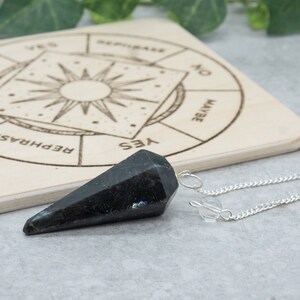 May include: A black pendulum with a silver chain on a wooden board with a circular chart that says "Yes", "No", "Maybe", and "Rephrase".