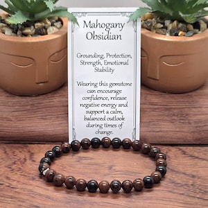 Peut inclure: Un bracelet composé de perles rondes en obsidienne acajou dans des tons marron et noir. Derrière le bracelet se trouve une carte rectangulaire blanche avec le texte Mahogany Obsidian, Grounding, Protection, Strength, Emotional Stability.