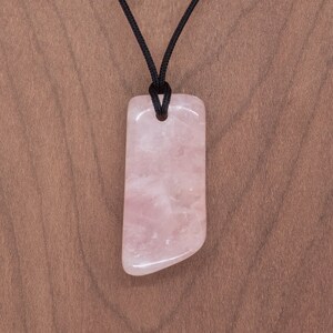 May include: A pink rose quartz pendant on a black cord necklace. The pendant is a smooth, polished, rectangular shape with rounded edges.