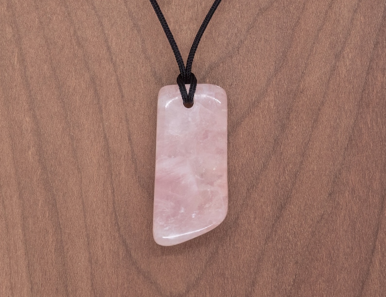 May include: A pink rose quartz pendant on a black cord necklace. The pendant is a smooth, polished, rectangular shape with rounded edges.