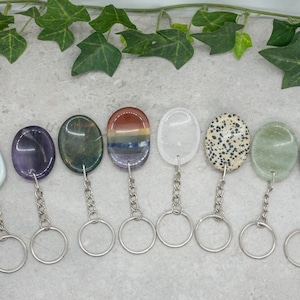 Op de afbeelding: Negen verschillende ovaalvormige edelsteen sleutelhangers met zilveren sleutelringen. De stenen zijn: lichtblauw, paars, groen, rood en blauw gestreept, wit, bruin en wit gevlekt, lichtgroen en grijs.