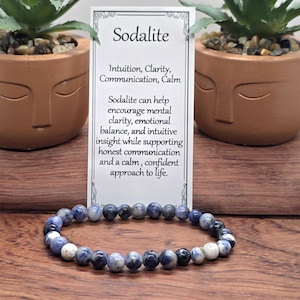 May include: A beaded bracelet made of round, polished sodalite stones in shades of blue, white, and dark grey. The bracelet is displayed in front of a white informational card titled Sodalite, which lists the properties of intuition, clarity, communication, and calm.