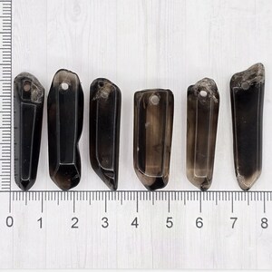May include: Six dark smoky quartz crystal pendants are arranged on a white surface next to a ruler. Each crystal is a different shape, with a hole drilled at the top for hanging. The crystals range from 1 to 2 inches in length.